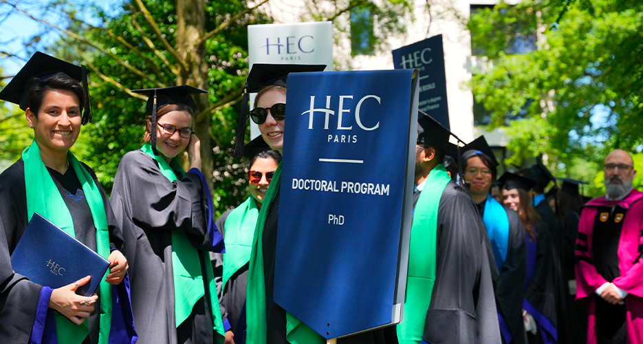 A group of HEC Paris doctoral graduates in academic regalia standing outdoors, holding a banner that reads 'HEC Paris Doctoral Program PhD'. The graduates are smiling and holding their diplomas, with several wearing sunglasses. The setting is a sunny outdoor area with trees and a building in the background. Additional HEC Paris banners are visible in the background