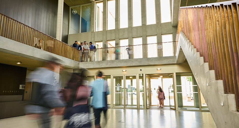 Intérieur d'un bâtiment universitaire moderne avec des étudiants marchant et se regroupant. L'espace spacieux est doté de grandes fenêtres, d'un escalier avec des garde-corps en bois et d'une disposition ouverte qui laisse entrer la lumière naturelle