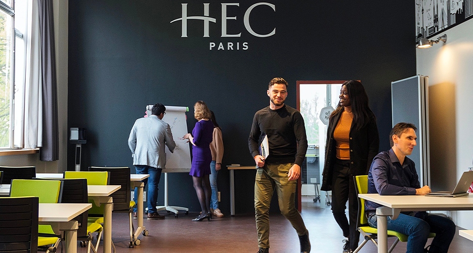 Étudiants interagissant dans une salle de classe moderne à HEC Paris. Certains discutent près d'un tableau blanc, tandis que d'autres travaillent sur des ordinateurs portables ou se déplacent dans la pièce