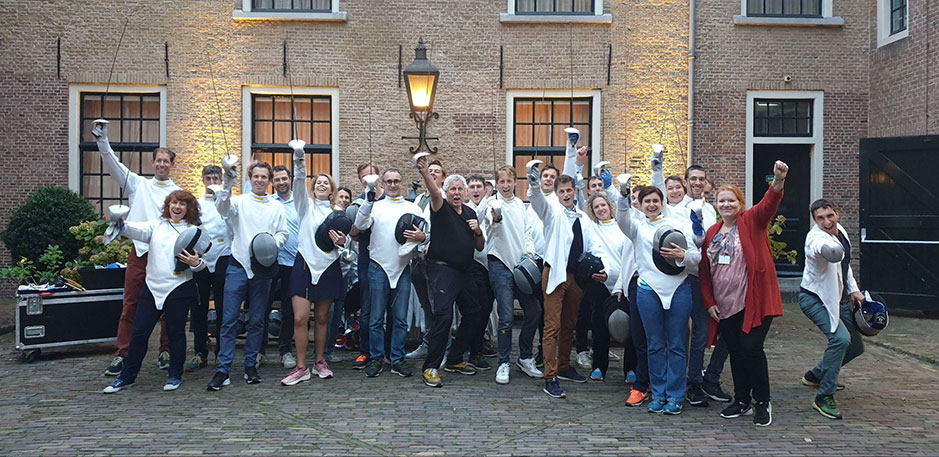 A group of people in fencing attire pose energetically in a courtyard, celebrating after a fencing session.