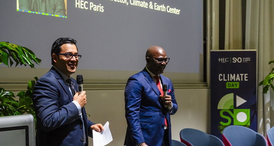 Fernando Diaz Lopez dressed in business attire, speak into microphones during the 'Climate Day' event at HEC Paris. They are standing in front of a projection screen with text and images, and next to a banner displaying the logos of HEC Paris and Climate and Earth, with the text 'Climate Day'.