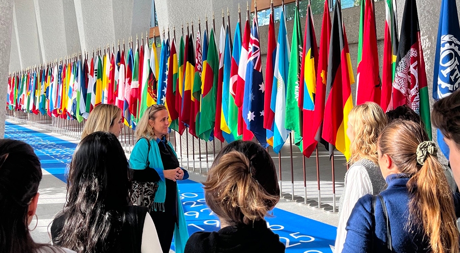 A group of people standing inside a building, facing a row of international flags from various countries