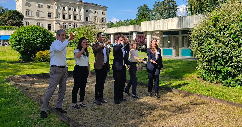 A group of six people standing outdoors on a grassy area, participating in an activity at HEC. They are casually dressed, with some wearing business casual attire, and appear to be engaged in a game, possibly throwing or aiming object