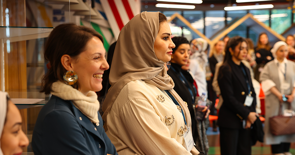 group of women attentively watching a presentation or event. One woman in the foreground is smiling and wearing a blue jacket with a white scarf, while another woman beside her, dressed in an embellished beige outfit with a beige hijab, looks on with a focused expression. Other women in various attire are visible in the background, creating a vibrant and multicultural scene in a warmly lit room with modern decor.