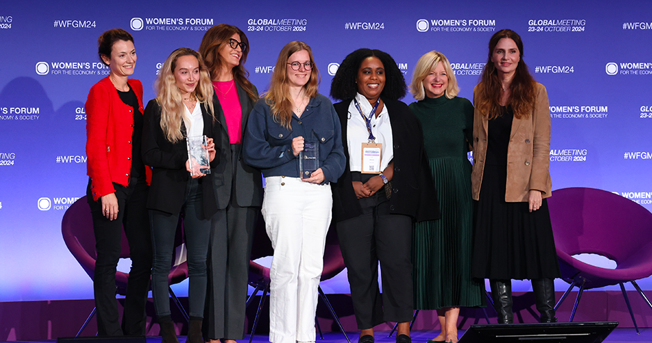 A group of seven women stand together on stage at the Women’s Forum for the Economy & Society, each smiling proudly. The background is a blue backdrop with the event’s logo and the date, 'Global Meeting 23-24 October 2024.' From left to right,