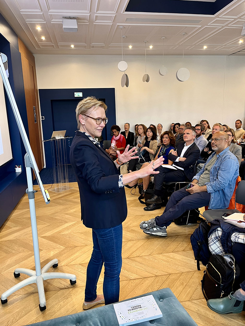 Une femme en veste sombre et jeans présente devant un tableau blanc dans une salle de conférence. Un public attentif est assis et écoute la présentation.