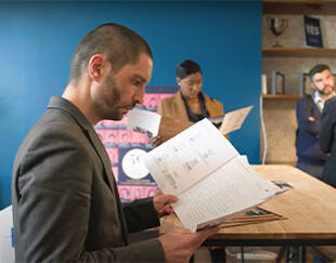 Un homme vêtu d'un costume regarde attentivement un document dans un espace de bureau, tandis que d'autres personnes en arrière-plan examinent également des papiers.