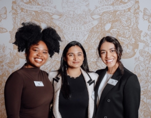 hree women smiling and standing together against a Christian Dior backdrop. The woman on the left has curly hair and is wearing a brown top. The woman in the middle has long black hair and is wearing a black top with a light-colored jacket. The woman on the right has long dark hair pulled back and is wearing a black blazer over a white top