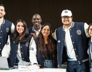 A group of six diverse students, wearing matching navy blue and white varsity jackets, smile at the camera. They are standing behind a table with a laptop and some papers, indicating they might be working on a project or event