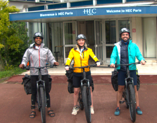 Three cyclists wearing sports gear and helmets pose with their electric bikes in front of the HEC Paris building entrance, under a sign that reads 'Welcome to HEC Paris'. They are dressed for a cycling adventure, ready to embark on an outdoor journey