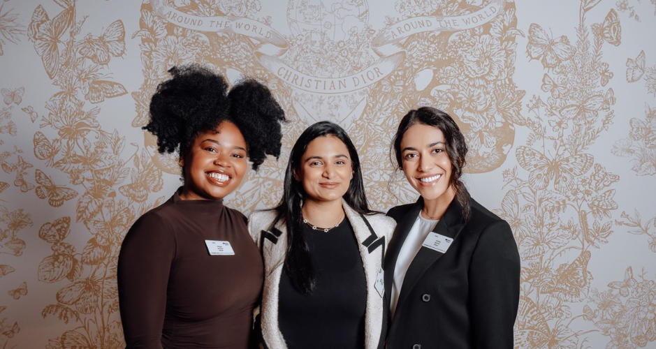 hree women smiling and standing together against a Christian Dior backdrop. The woman on the left has curly hair and is wearing a brown top. The woman in the middle has long black hair and is wearing a black top with a light-colored jacket. The woman on the right has long dark hair pulled back and is wearing a black blazer over a white top