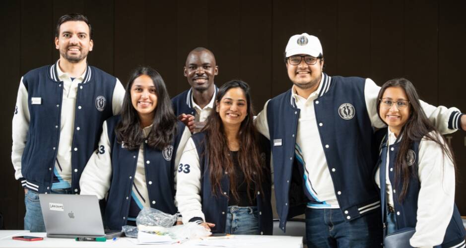 A group of six diverse students, wearing matching navy blue and white varsity jackets, smile at the camera. They are standing behind a table with a laptop and some papers, indicating they might be working on a project or event