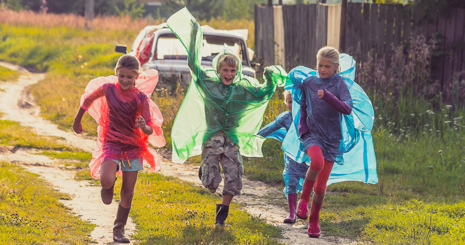 Four children joyfully running on a dirt path, wearing colorful transparent plastic capes. The children are having fun outdoors, with bright smiles on their faces