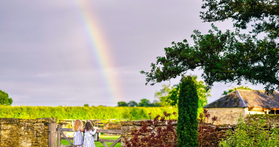 Two young girls in white dresses stand by a wooden gate, gazing at a vibrant rainbow stretching across a cloudy sky