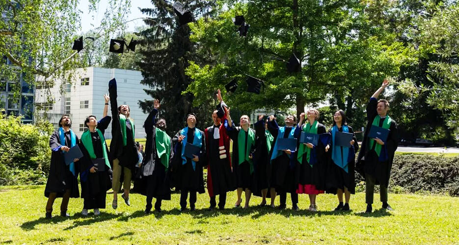A group of doctoral candidates in graduation gowns and hoods celebrating outdoors by throwing their caps into the air. They are standing on a grassy area surrounded by trees and a building in the background. The graduates are smiling and holding their diplomas as they commemorate this significant milestone.
