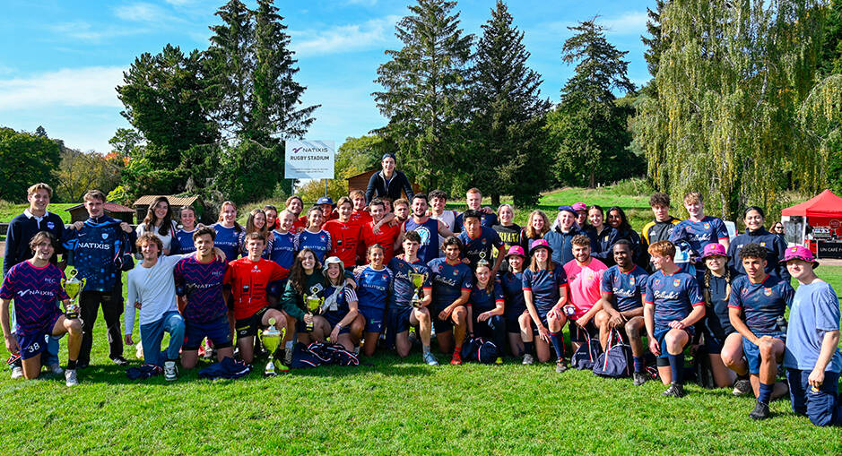 Photo de groupe des équipes gagnantes de la Natixis Rugby Cup posant sur le terrain avec leurs trophées, entourées de nature et d'arbres en arrière-plan