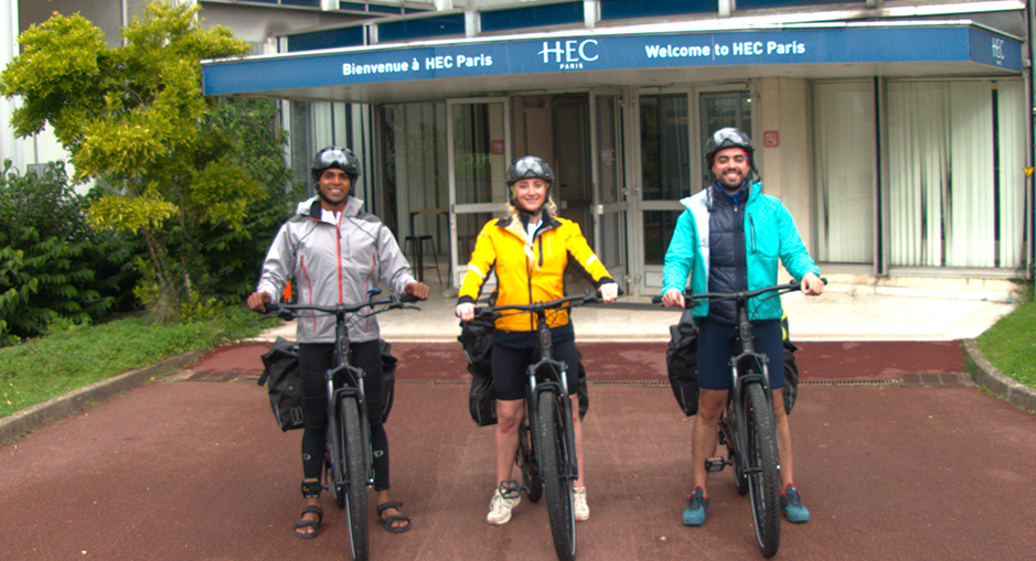 Three cyclists wearing sports gear and helmets pose with their electric bikes in front of the HEC Paris building entrance, under a sign that reads 'Welcome to HEC Paris'. They are dressed for a cycling adventure, ready to embark on an outdoor journey