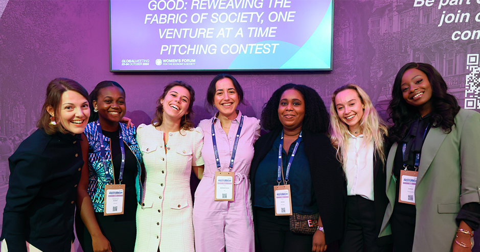 seven smiling women pose together at the Women’s Forum event. They are dressed in professional attire and wear accreditation badges around their necks. Behind them, a purple backdrop features a large screen with the text 'GOOD: Reweaving the Fabric of Society, One Venture at a Time Pitching Contest.' The women are standing close to each other, conveying a sense of camaraderie and positivity