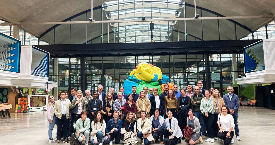 Group of executives and professionals gathered at a modern innovation hub, with a colorful sculpture and industrial-style architecture in the background